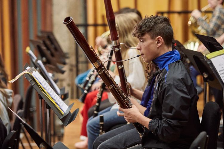 A boy wearing a grey jacket with blue collar sits playing a bassoon. Other people are sat around him.
